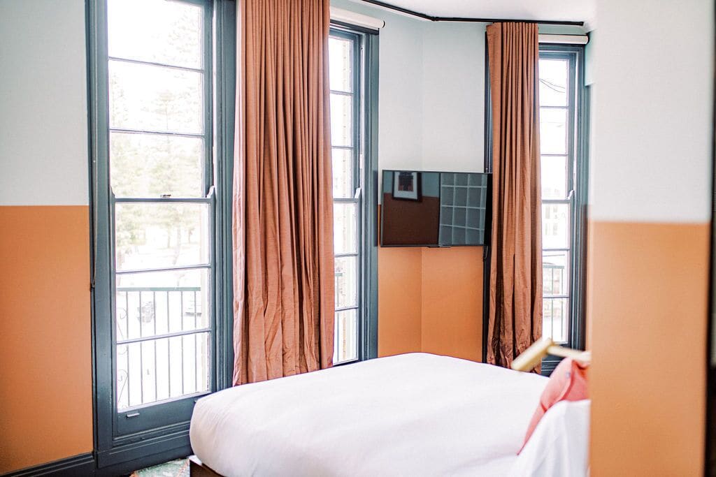 Habitación de hotel con cama, dos ventanas con cortinas, aire acondicionado de pared y vistas al exterior.