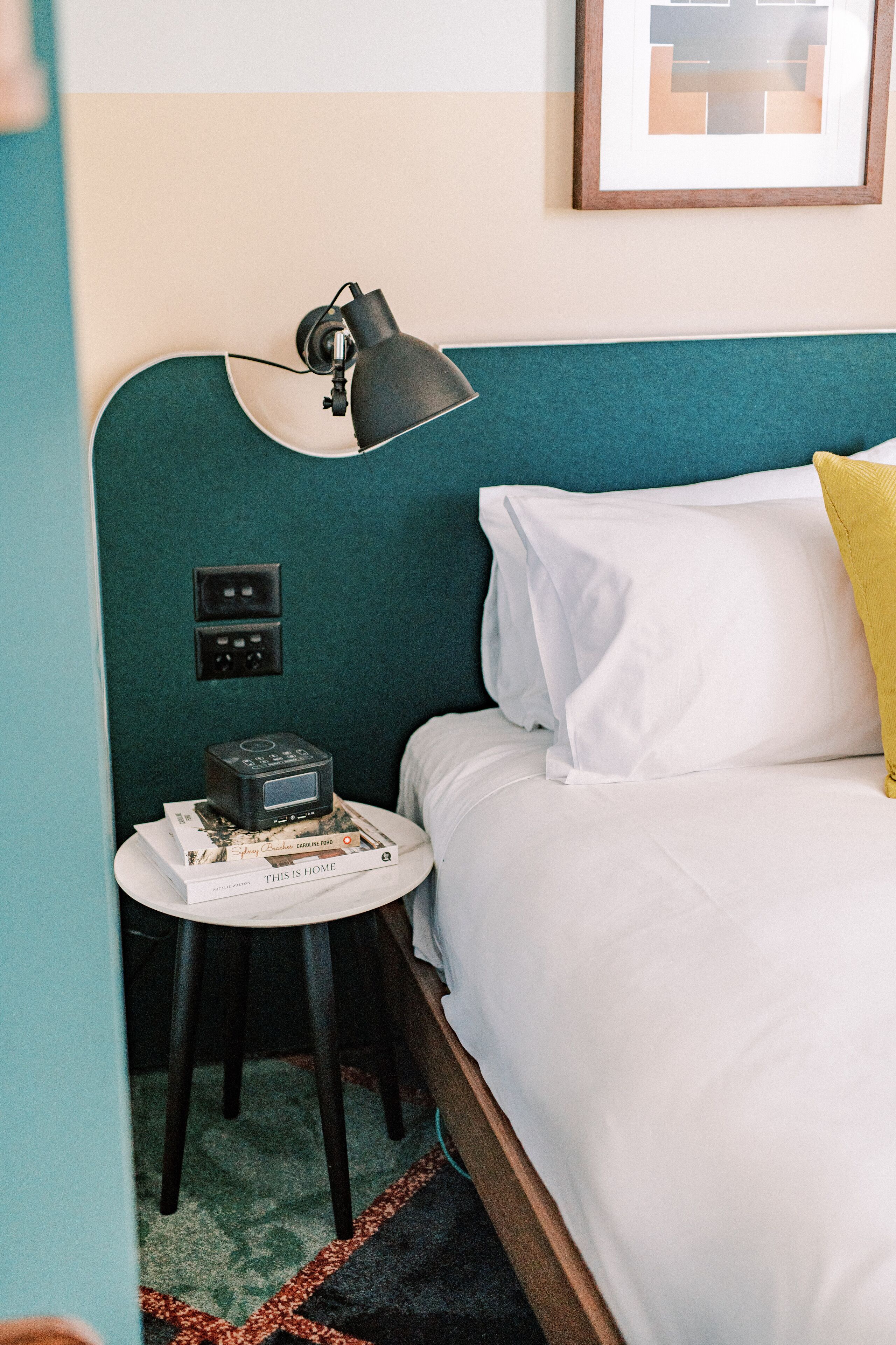 A neatly made bed with white and yellow pillows, a small bedside table with a lamp, and a wall-mounted power outlet.