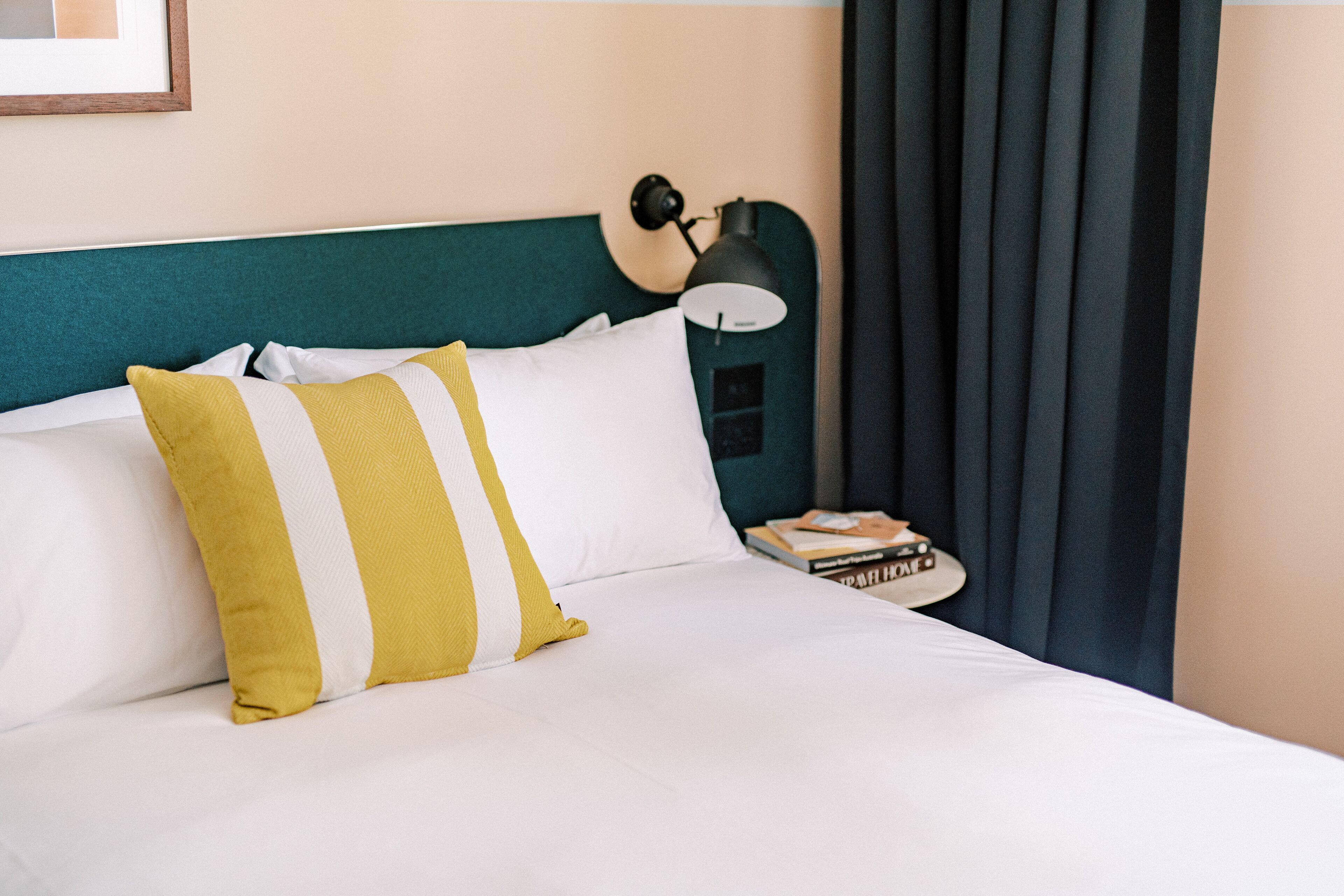 A neatly made bed with white and yellow striped pillows, a teal headboard, a black lamp, and a stack of books on a bedside table.