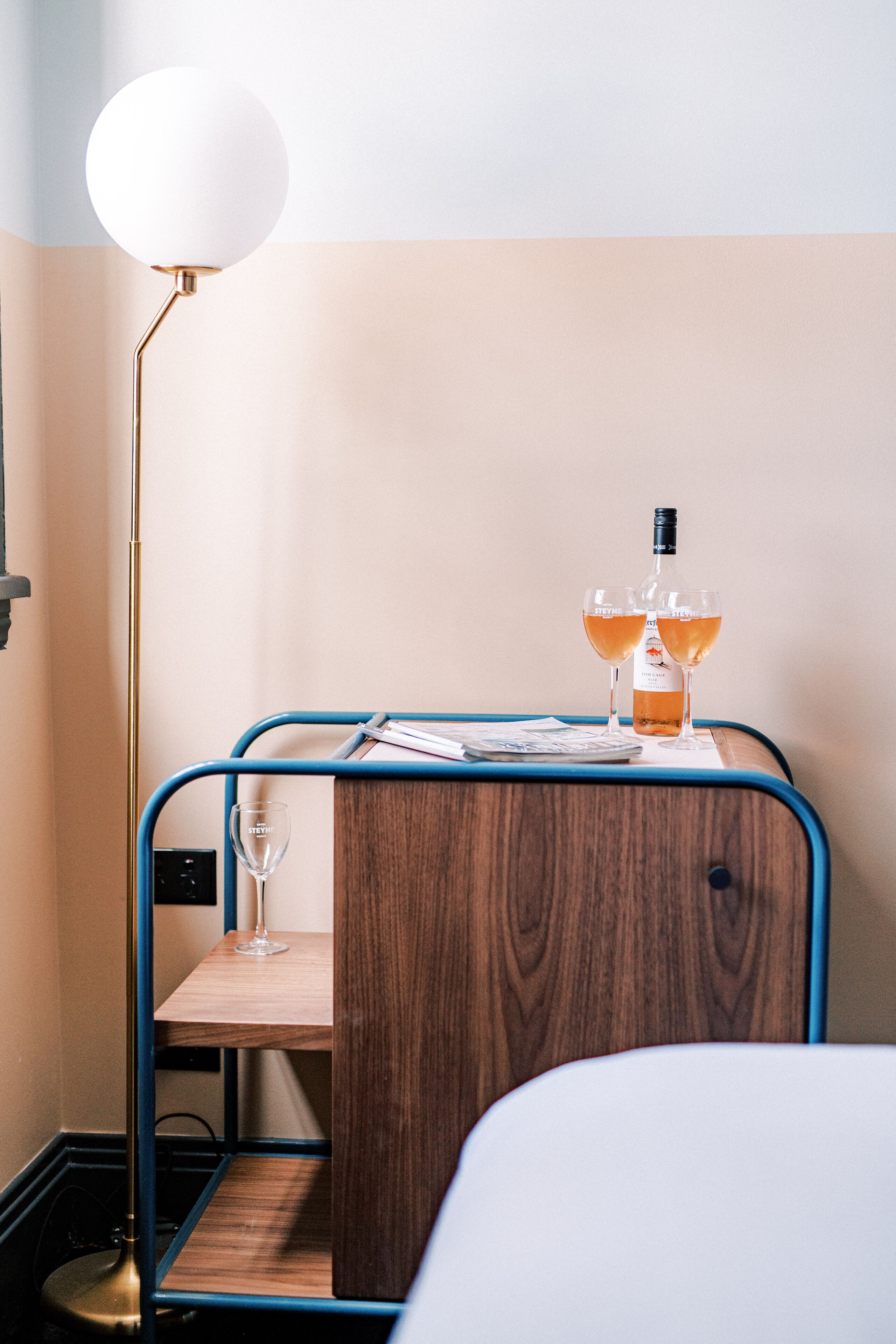 A small wooden table with a bottle and two glasses of wine, a floor lamp, and a bed in the background.