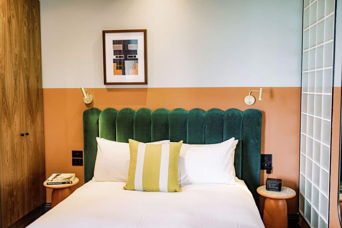 A hotel room with a green upholstered headboard, white bedding, a framed artwork on the wall, a bedside table with a lamp, and a small table with a radio.