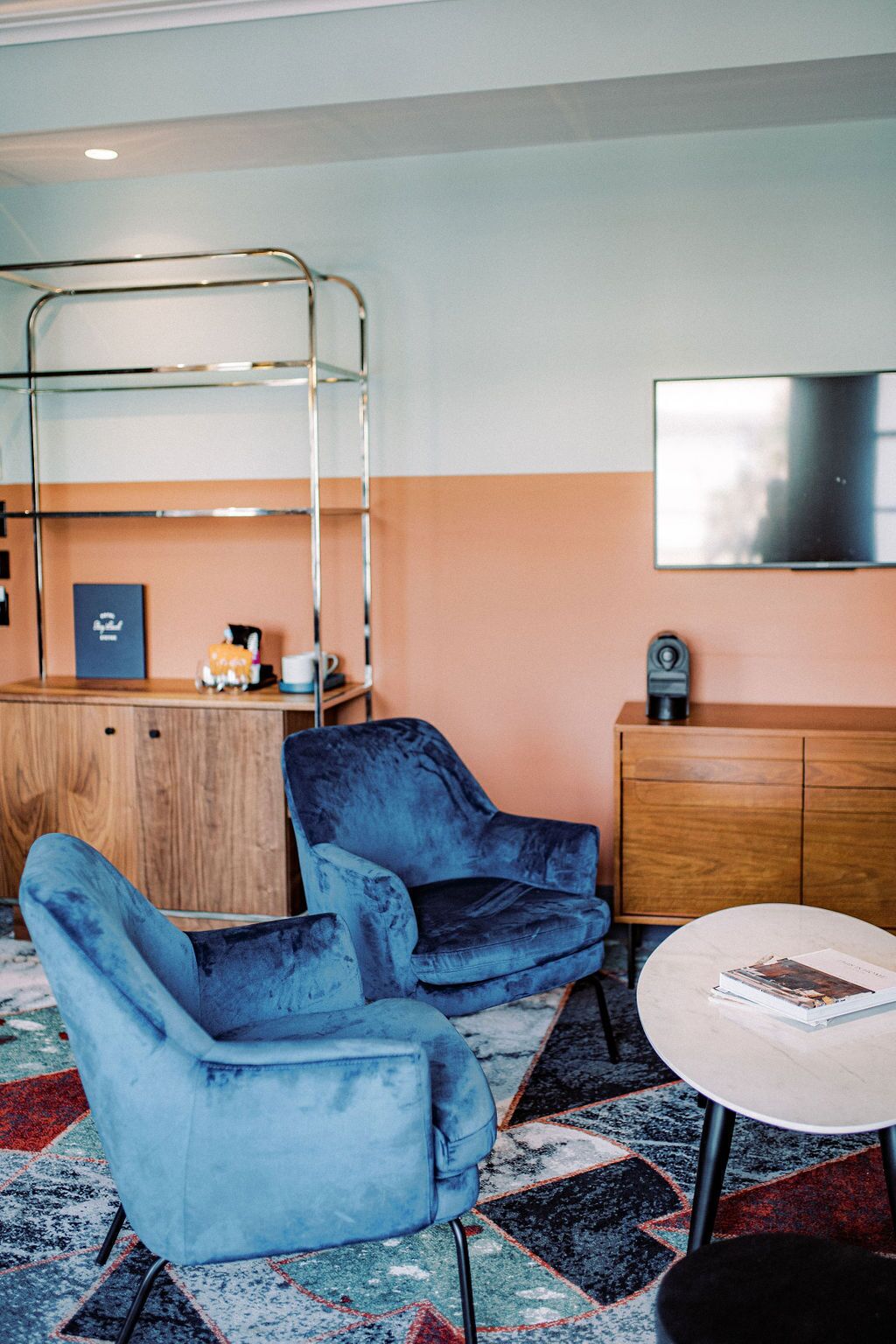 Habitación de hotel con una cama, dos sillones azules, una mesita redonda, un armario de madera, un televisor y una alfombra estampada.
