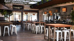 Un restaurante interior con mesas de madera y taburetes de bar blancos, una zona de bar con estantes llenos de botellas y plantas colgantes.