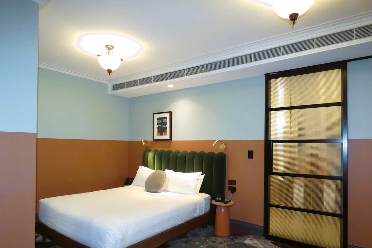 A hotel room with a bed, a nightstand, a wall-mounted clock, a framed picture, and a window with blinds.