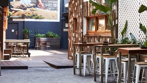 Un patio con mesas y sillas de madera, plantas en macetas y un mural grande en la pared.
