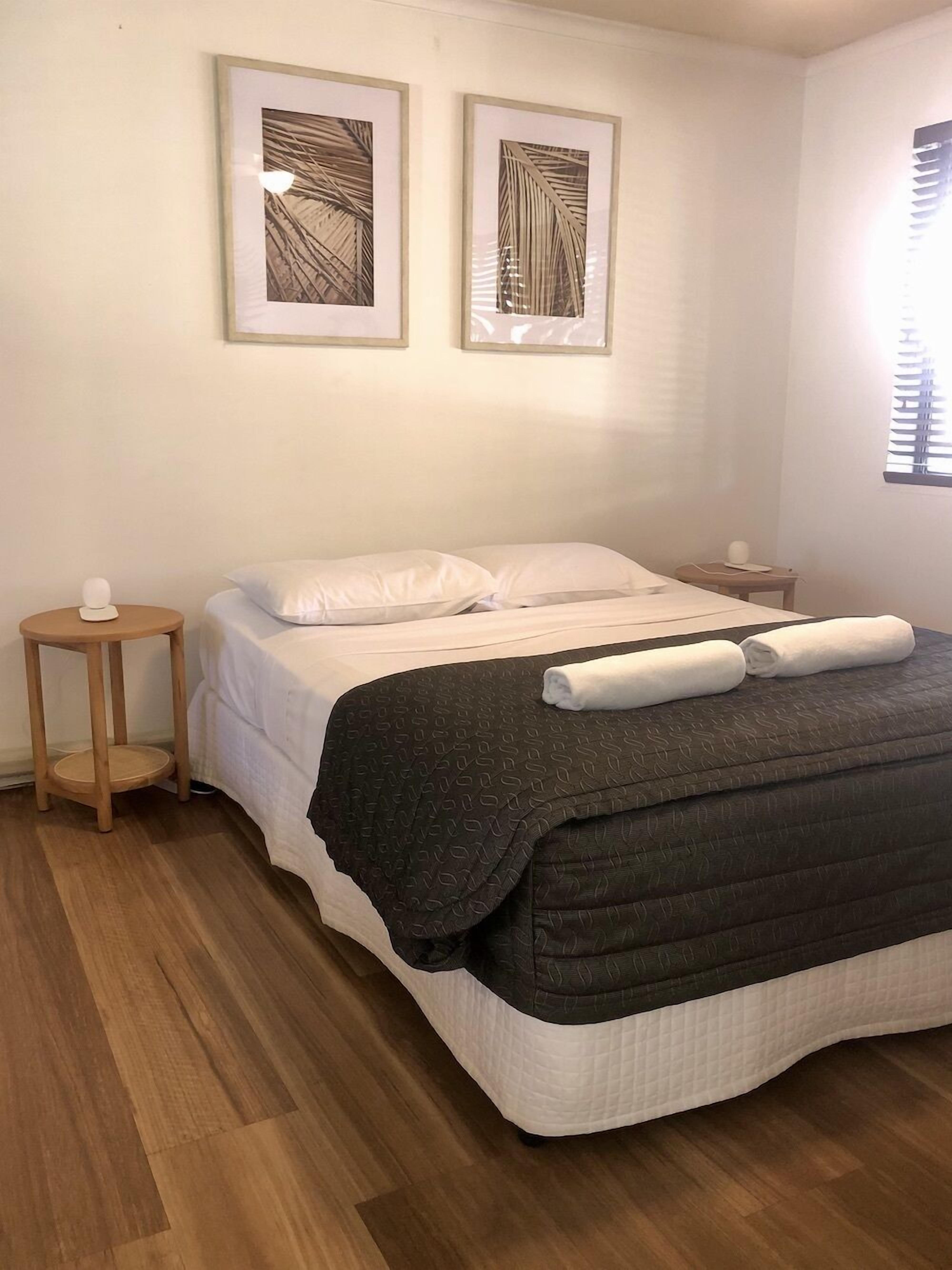 Un lit bien fait, recouvert d’une couvre-lit à motifs sombres, deux œuvres d’art abstraites encadrées accrochées au mur et un plancher en bois.
