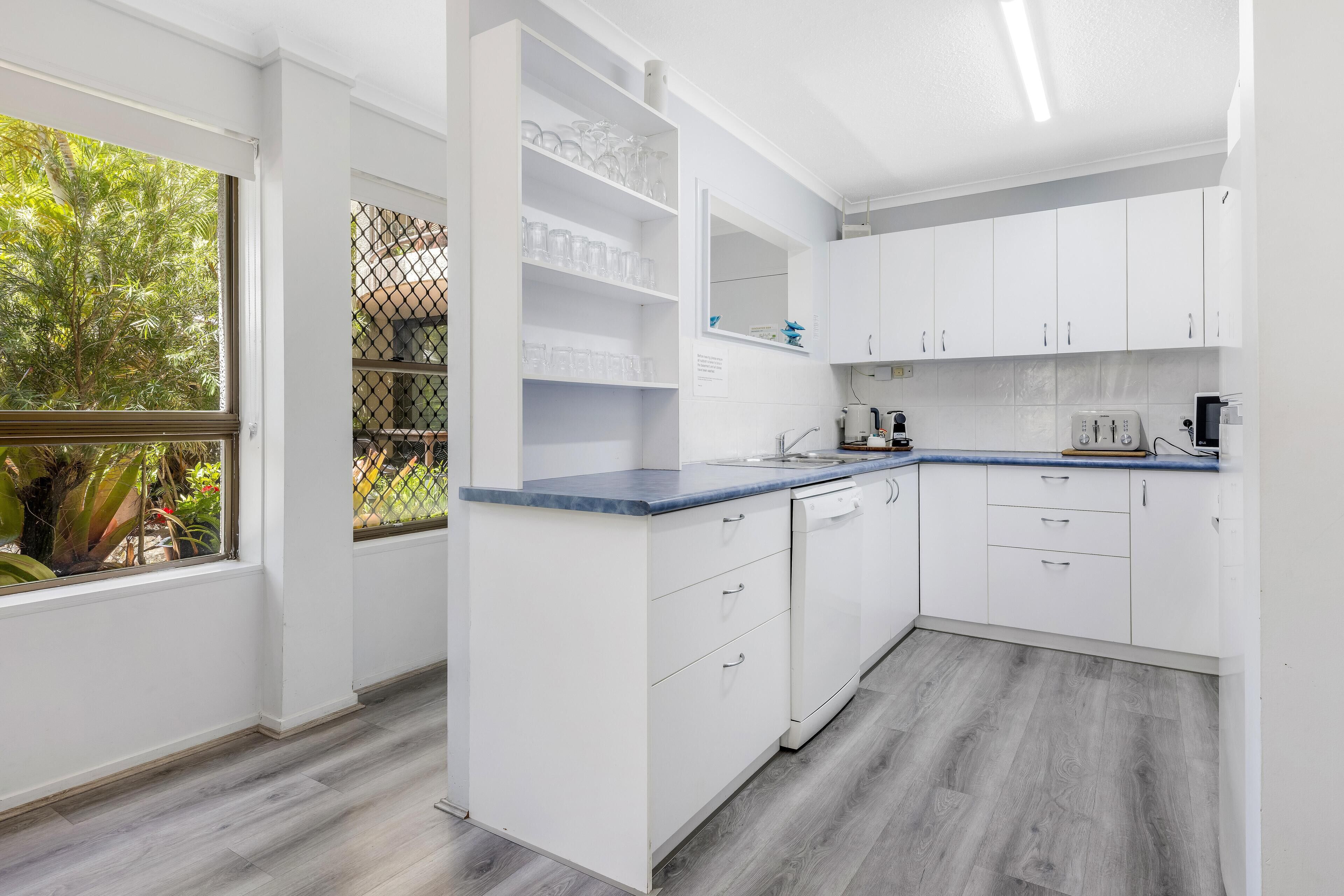Une cuisine moderne avec des meubles blancs, un plan de travail bleu et une fenêtre donnant sur un paysage verdoyant.