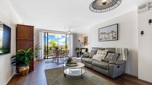 A modern living room with a grey sofa, a coffee table, and a large window offering a view of palm trees. - Kalua Holiday Apartments (Maroochydore)