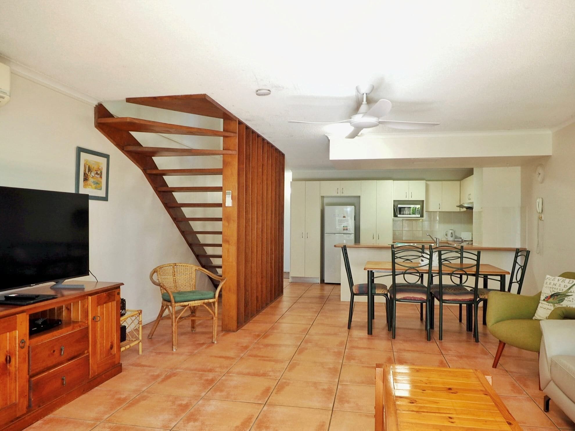 A living room with a TV, wooden entertainment unit, wicker chair, dining table with chairs, and a staircase.