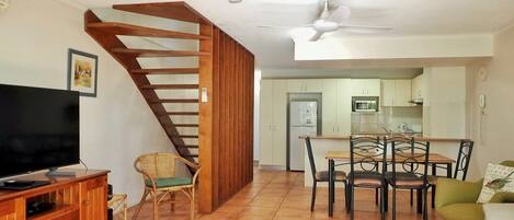 A living room with a TV, wooden entertainment unit, wicker chair, dining table with chairs, and a staircase.
