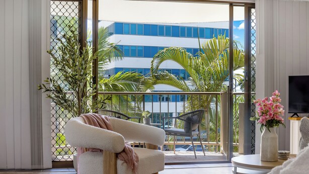 A modern living room with a white sofa, a coffee table, and a TV. A balcony with plants and a building is visible through the glass doors. - Kalua Holiday Apartments (Maroochydore)
