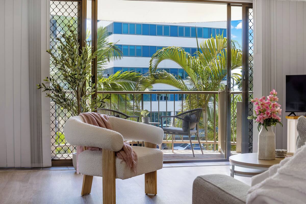 A modern living room with a white sofa, a coffee table, and a TV. A balcony with plants and a building is visible through the glass doors.