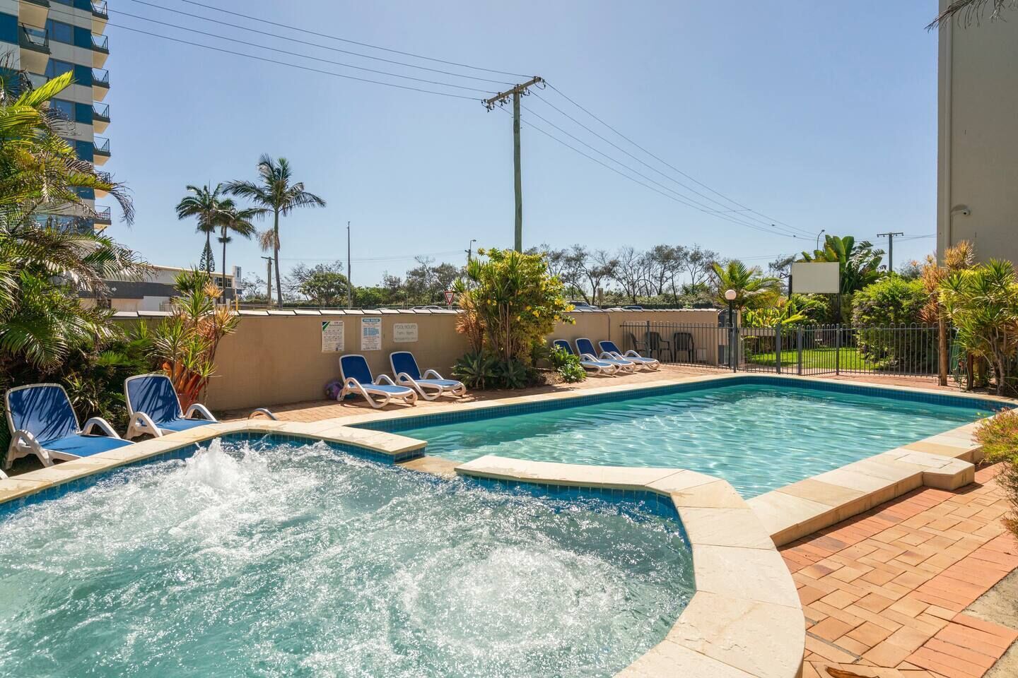 Una piscina con sillas de descanso y un jacuzzi, rodeada de palmeras y un edificio.