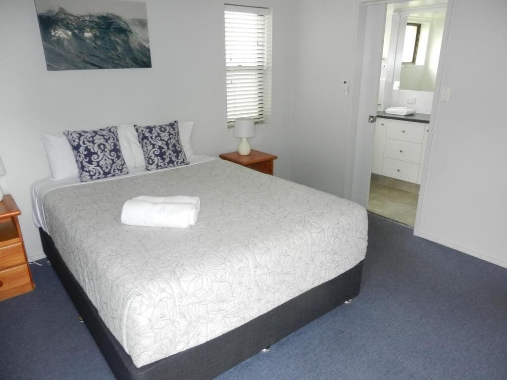 Una cama bien hecha con edredón blanco y dos almohadas estampadas, una mesita de noche con una lámpara y un cuadro en la pared.