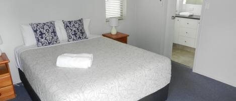 Una cama bien hecha con edredón blanco y dos almohadas estampadas, una mesita de noche con una lámpara y un cuadro en la pared.