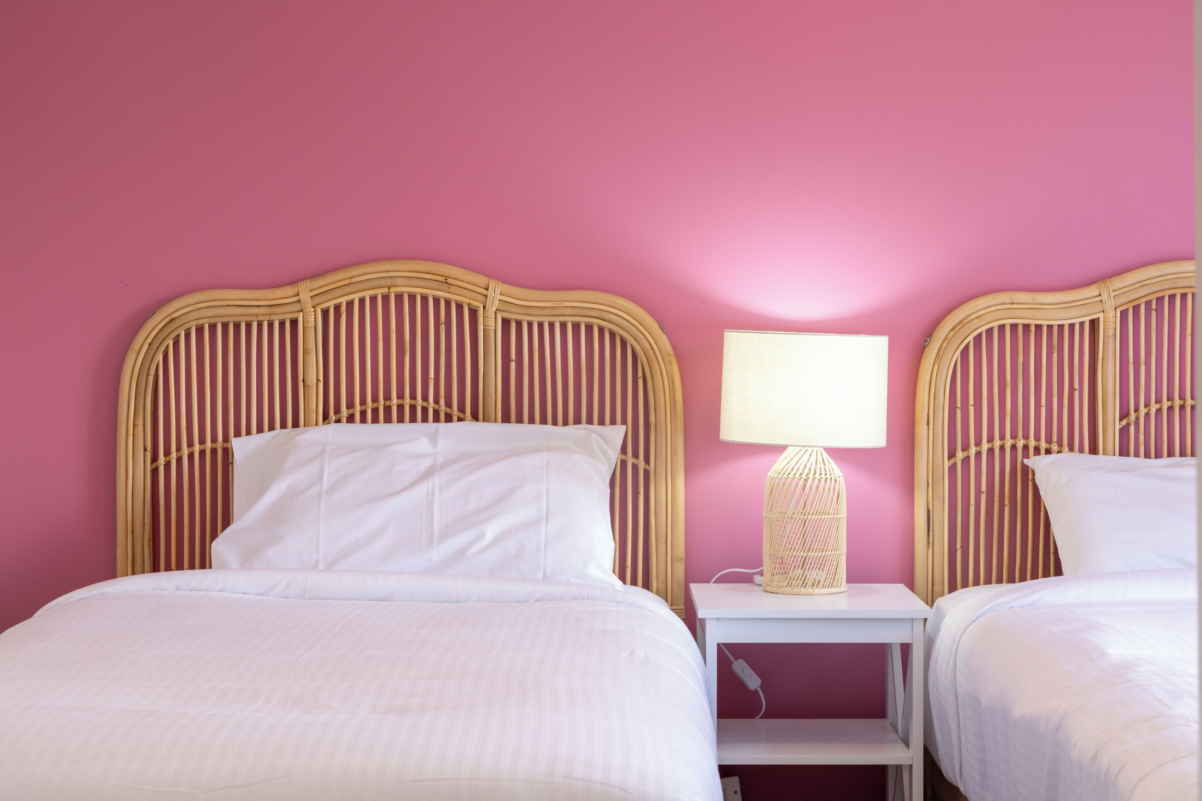 A room with two beds, each with a wicker headboard, a bedside table with a lamp, and a pink wall.