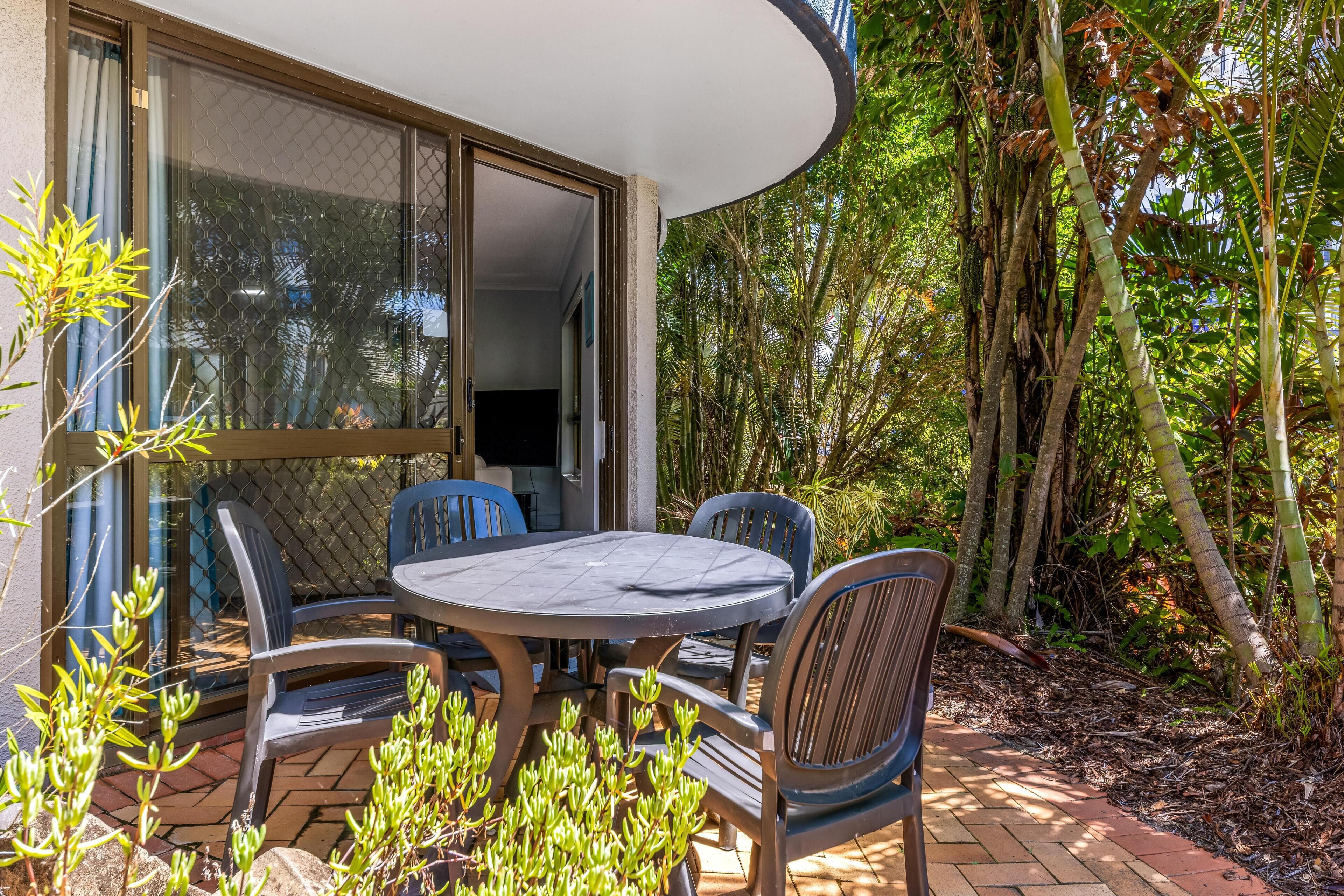 An outdoor patio with a round table and chairs, surrounded by lush greenery and a large window overlooking the garden.