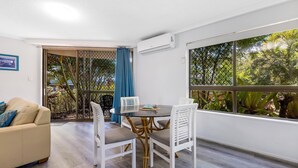 A modern room with a glass table, white chairs, a beige sofa, and large windows with a view of greenery. - Kalua Holiday Apartments (Maroochydore)
