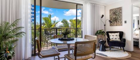 A modern living room with a round table, chairs, a black armchair, and a view of palm trees through large sliding doors.
