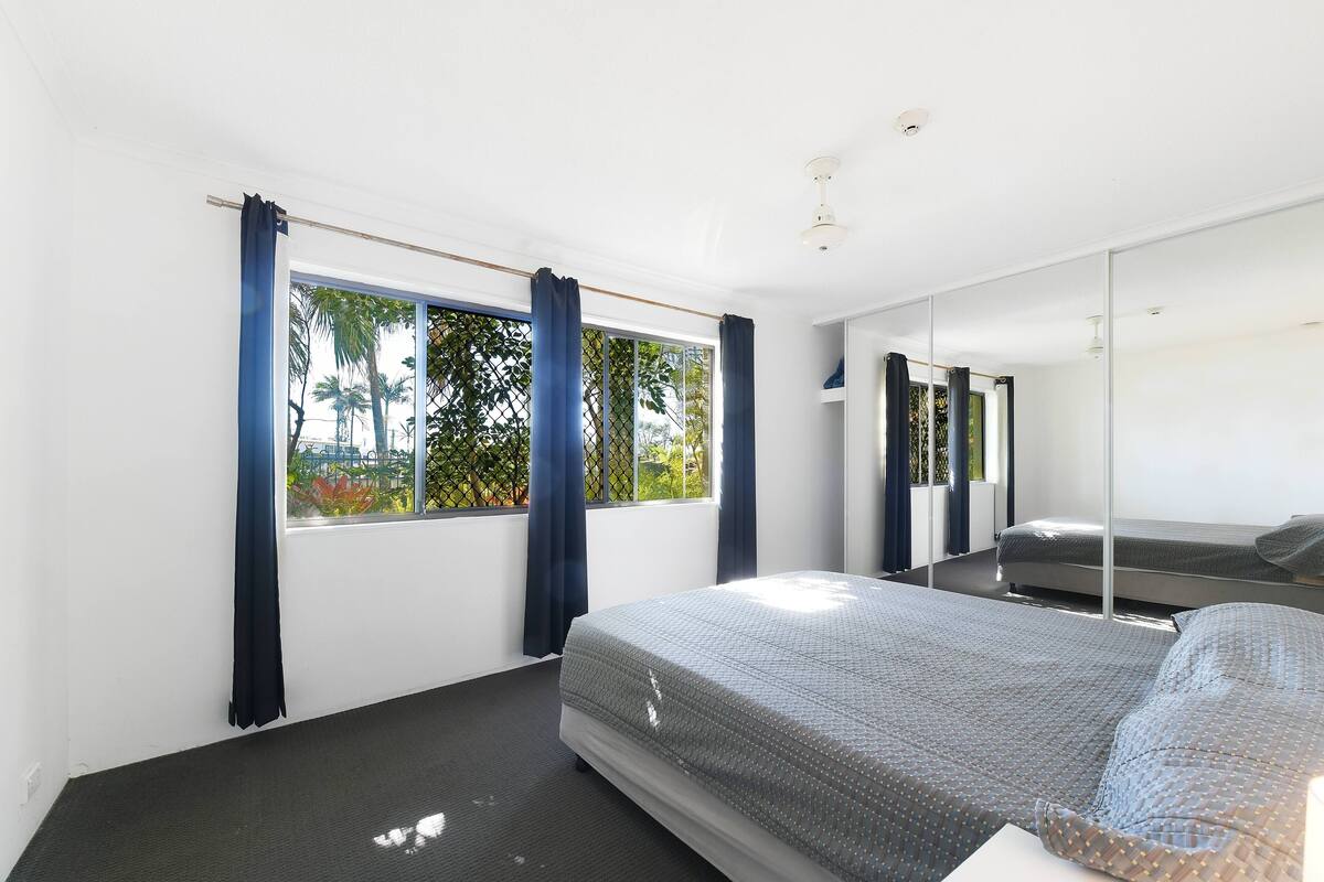A modern bedroom with a large bed, a window with curtains, a mirror, and a view of greenery outside.