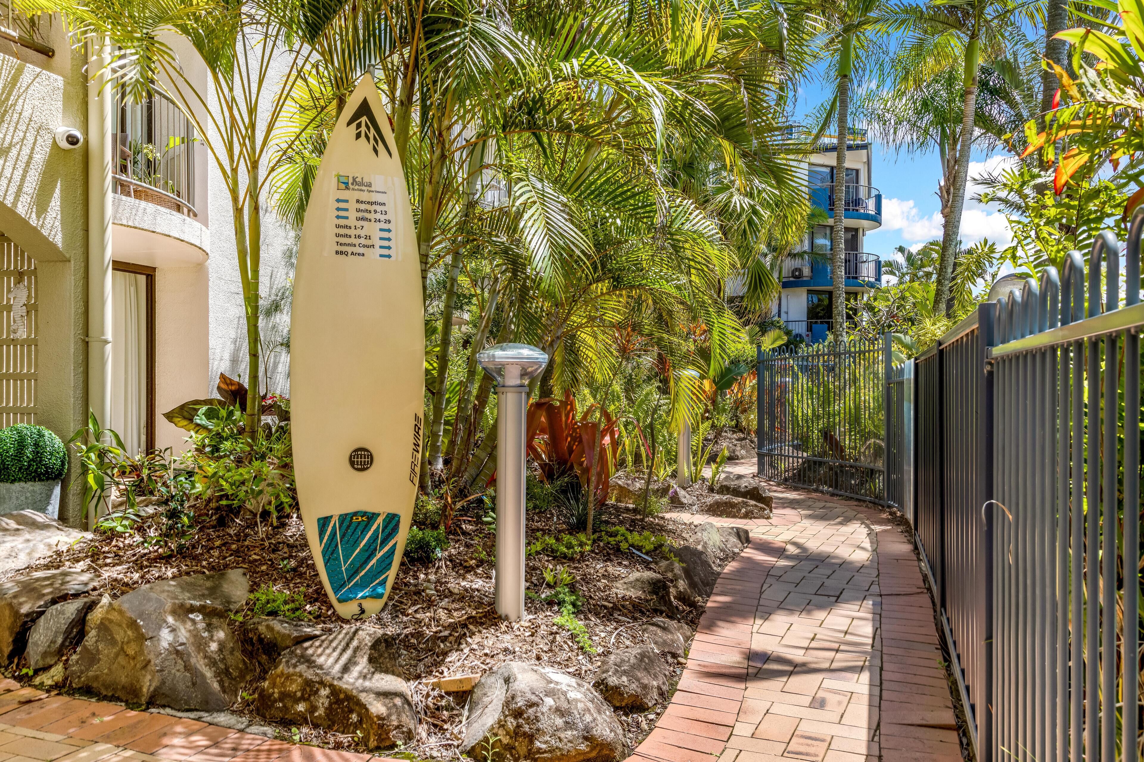 Un sendero bordeado por plantas tropicales y un cartel con una tabla de surf que indica los servicios del hotel.