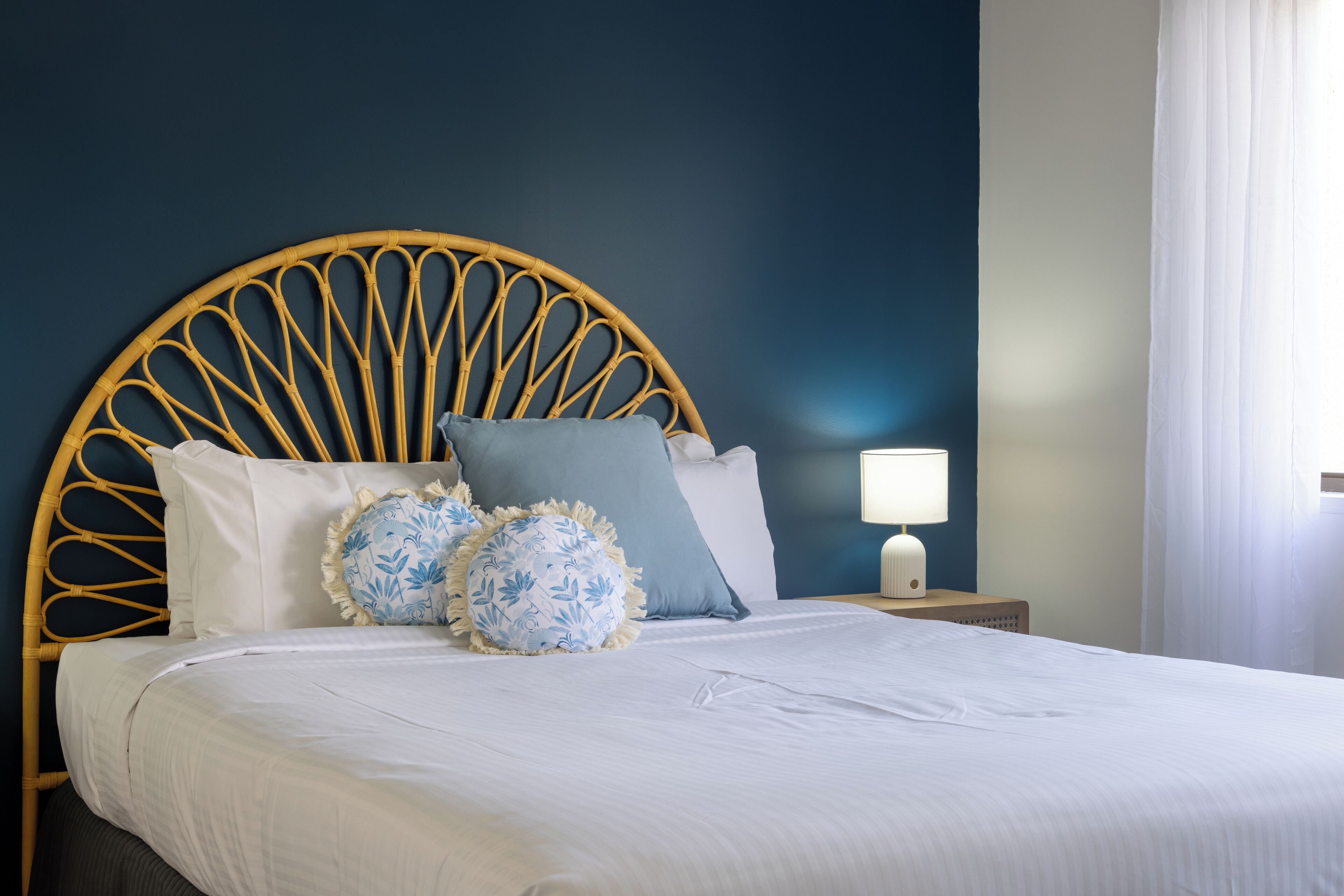 Una cama bien hecha con cabecera de ratán, ropa de cama blanca y dos almohadas decorativas.