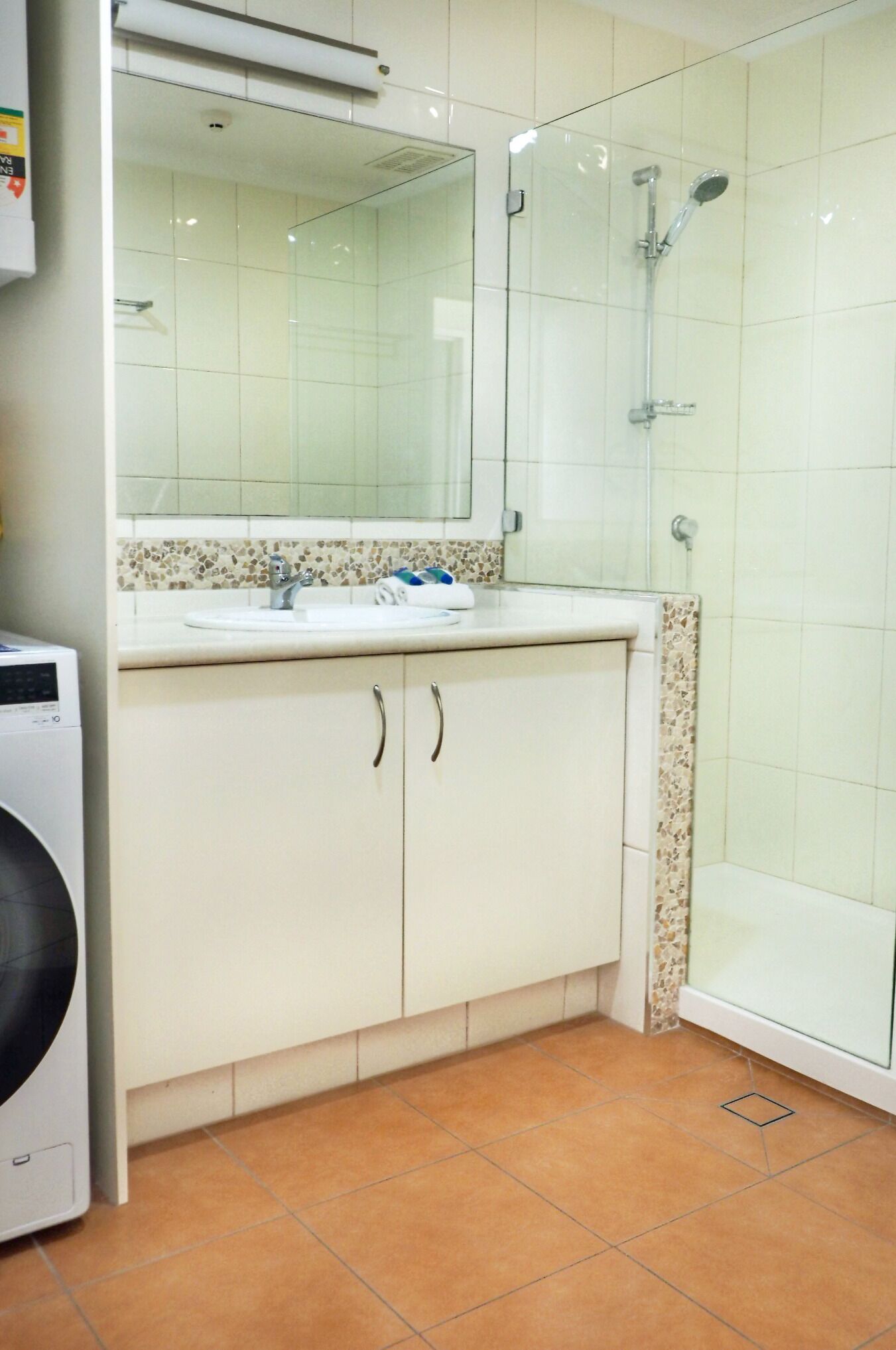 A bathroom with a glass shower enclosure, a sink with a mirror above it, and a washing machine to the left.