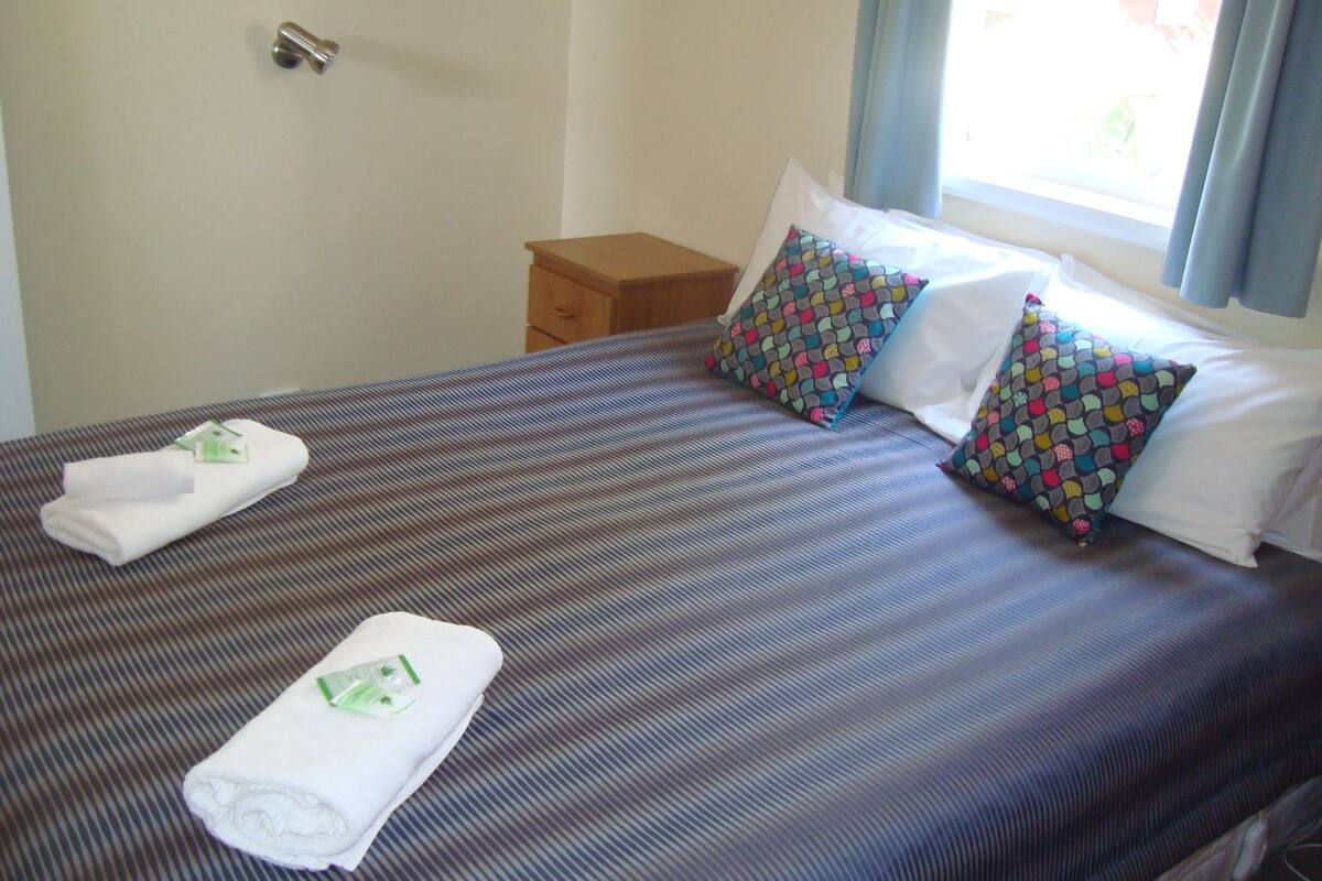 A hotel room with a bed, two pillows, a nightstand, and a window with curtains.