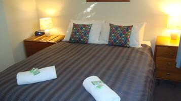 Una habitación de hotel con una cama bien hecha, dos lámparas de noche, una mesita de noche de madera y un cuadro enmarcado en la pared.