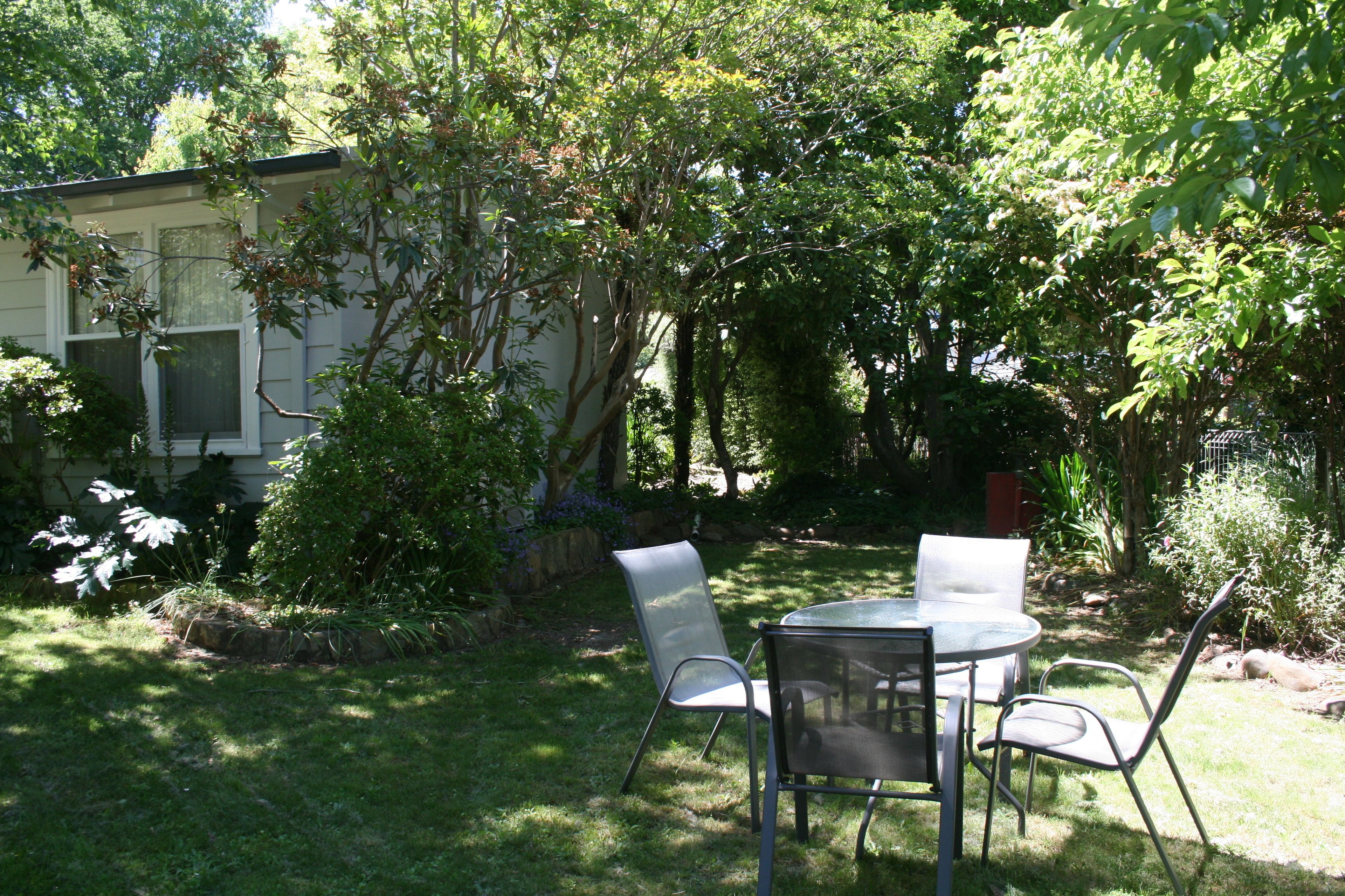 A patio with a round table and four chairs, surrounded by lush greenery and a house in the background.