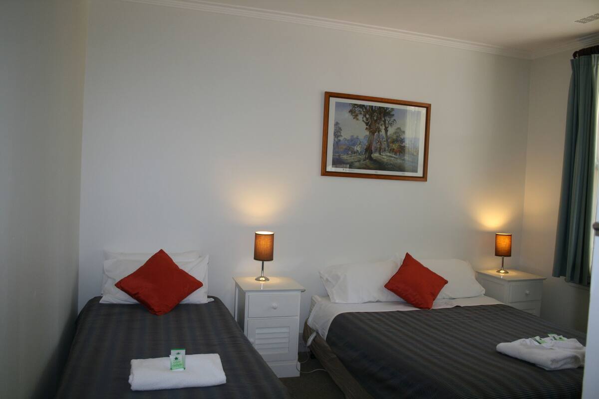A hotel room with two beds, each with a red pillow, white linens, and a small bedside table with a lamp. There is a framed painting on the wall above the beds.