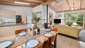 Cozinha moderna com armários de madeira, sala de jantar com mesa posta para quatro e sala de estar com sofá e televisão.