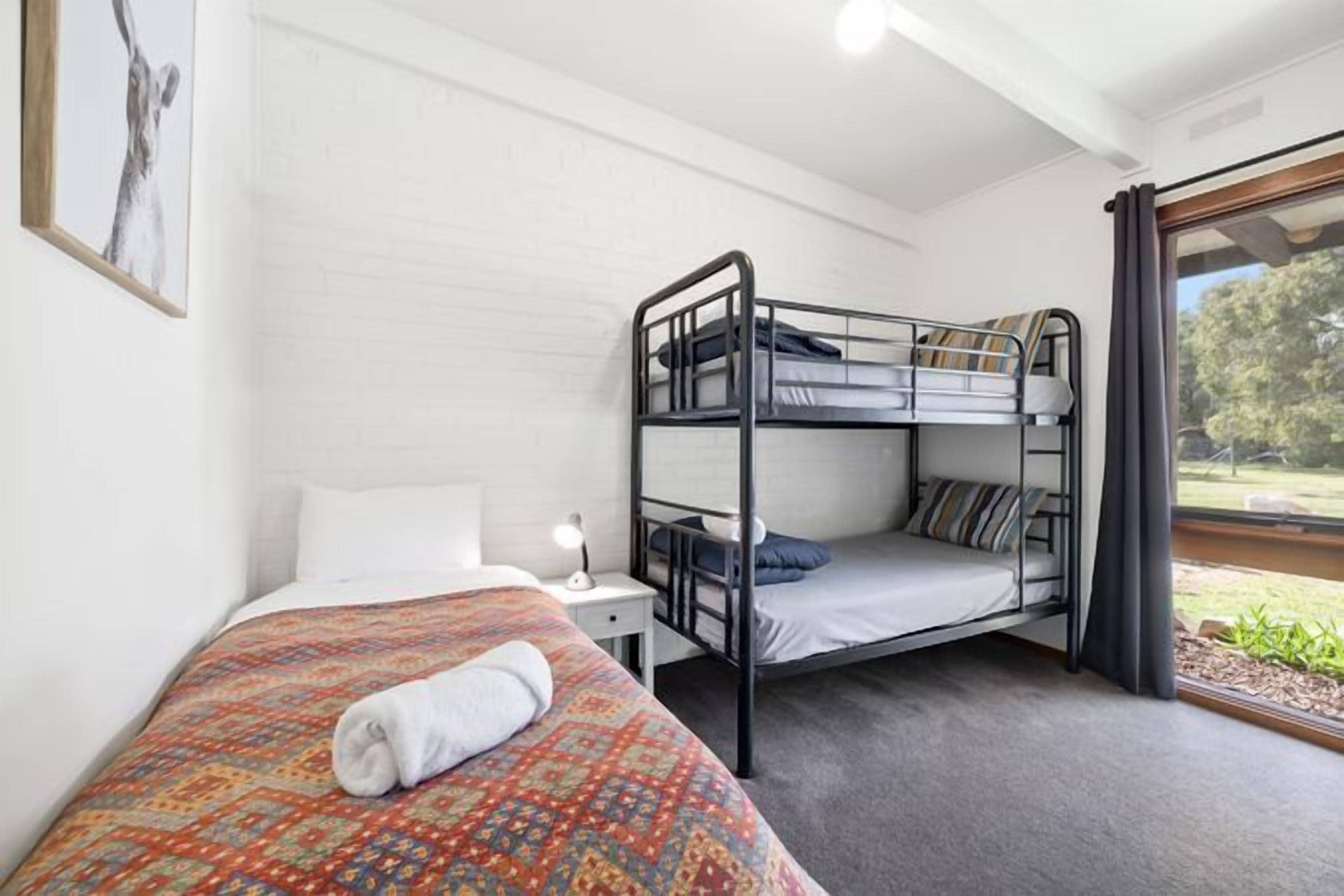 A room with a bunk bed, a single bed, a nightstand with a lamp, and a window with curtains.