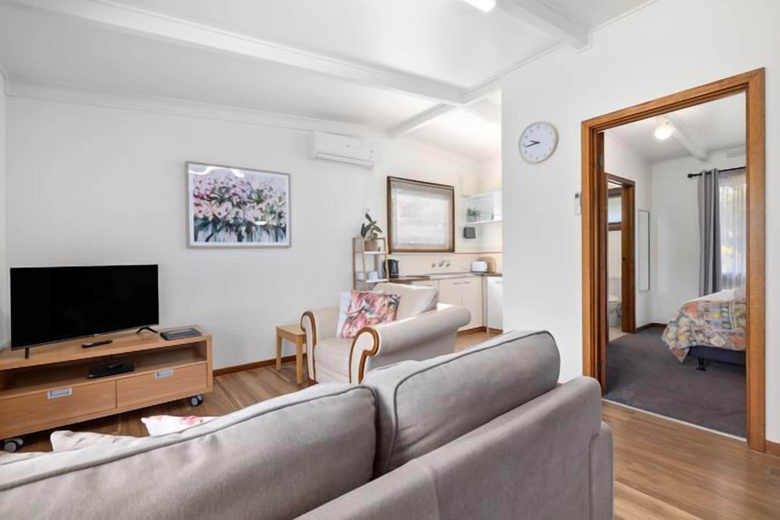 Ein modernes Wohnzimmer mit Fernseher auf einem Holzständer, einem grauen Sofa, einem kleinen Tisch und einem Gemälde an der Wand.