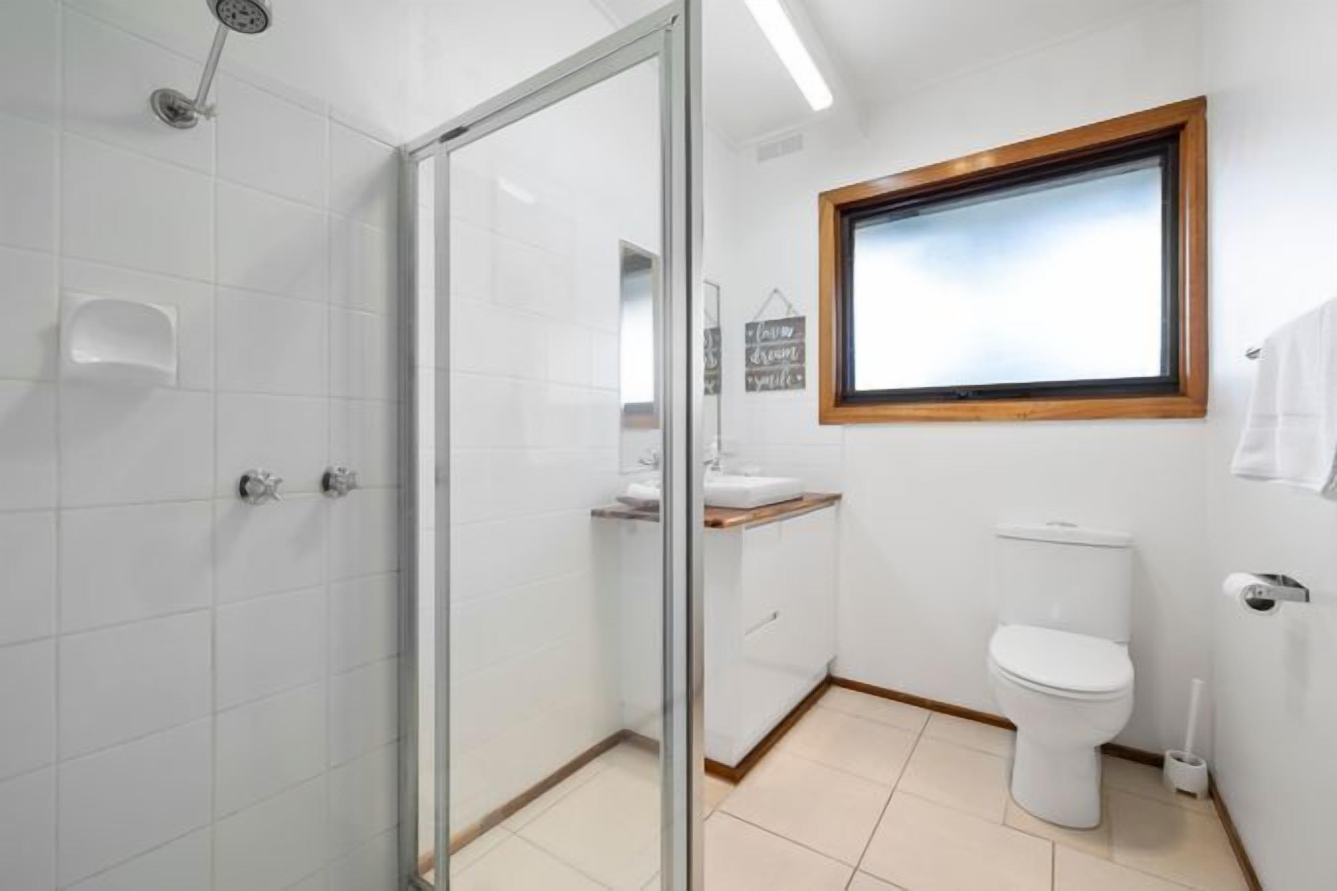 A bathroom with a glass shower enclosure, a white toilet, a sink with a wooden countertop, and a window with a wooden frame.