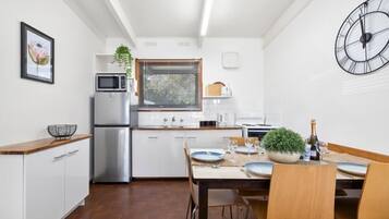 Cocina compacta con armarios blancos, microondas y refrigerador. Hay una mesa de comedor con sillas y un reloj en la pared.