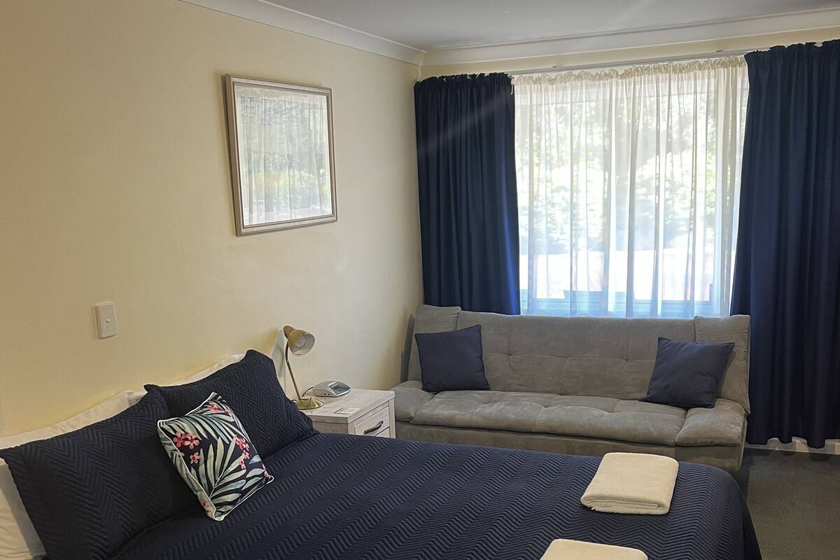 A hotel room with a large bed, a sofa, a bedside table with a lamp, and a window with curtains.
