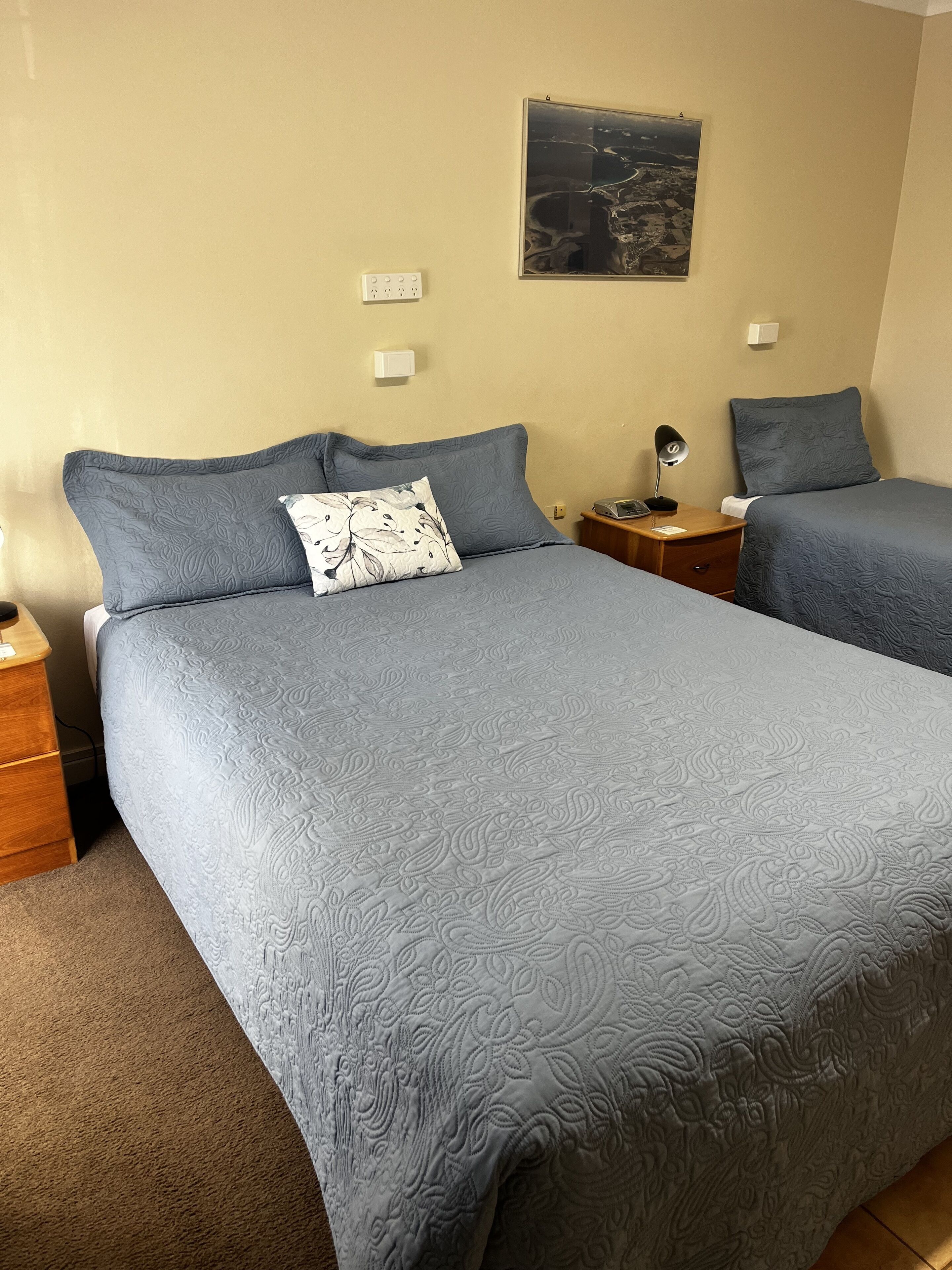 A double bed with a floral-patterned pillow, a nightstand with a lamp, and a wall-mounted power outlet.