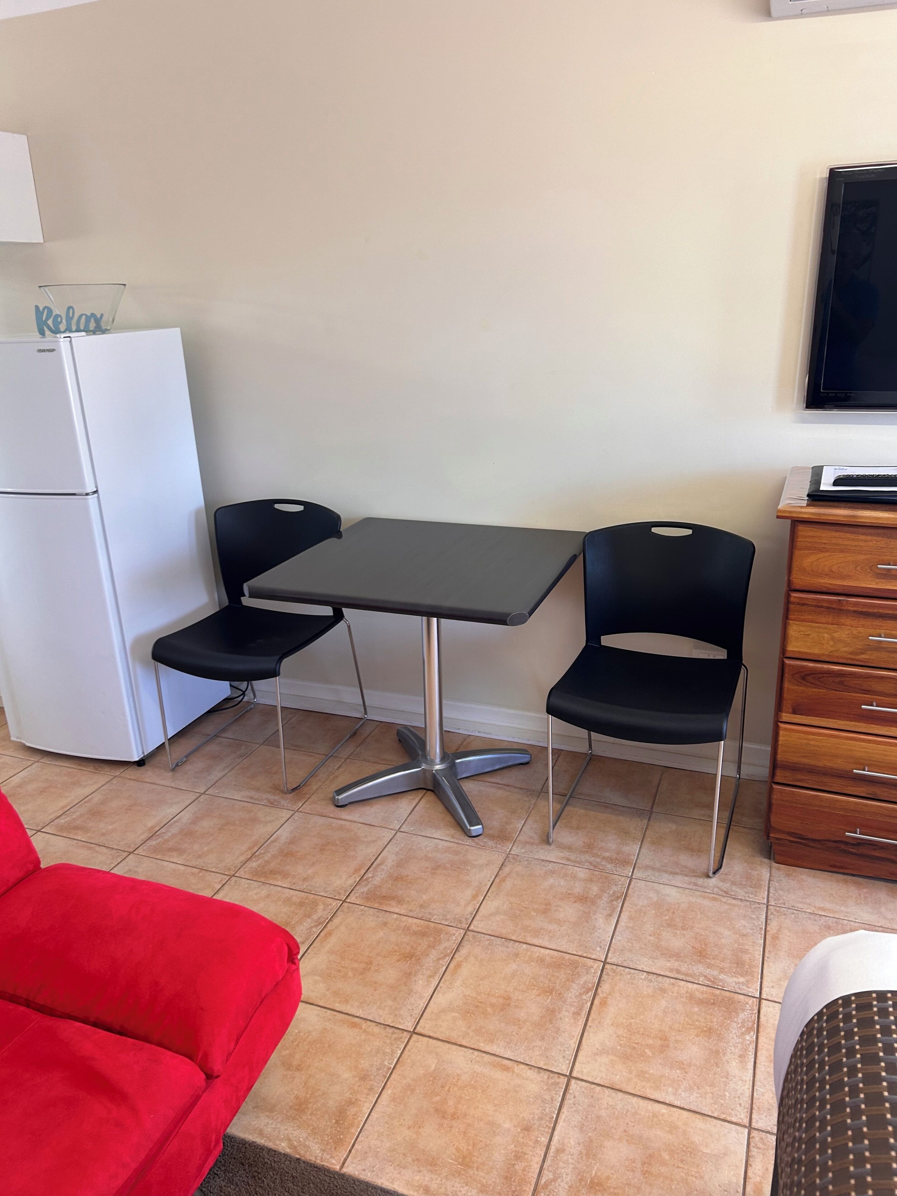 Una habitación compacta con kitchenette, una mesa de comedor con dos sillas, un sofá rojo y un televisor en la pared.