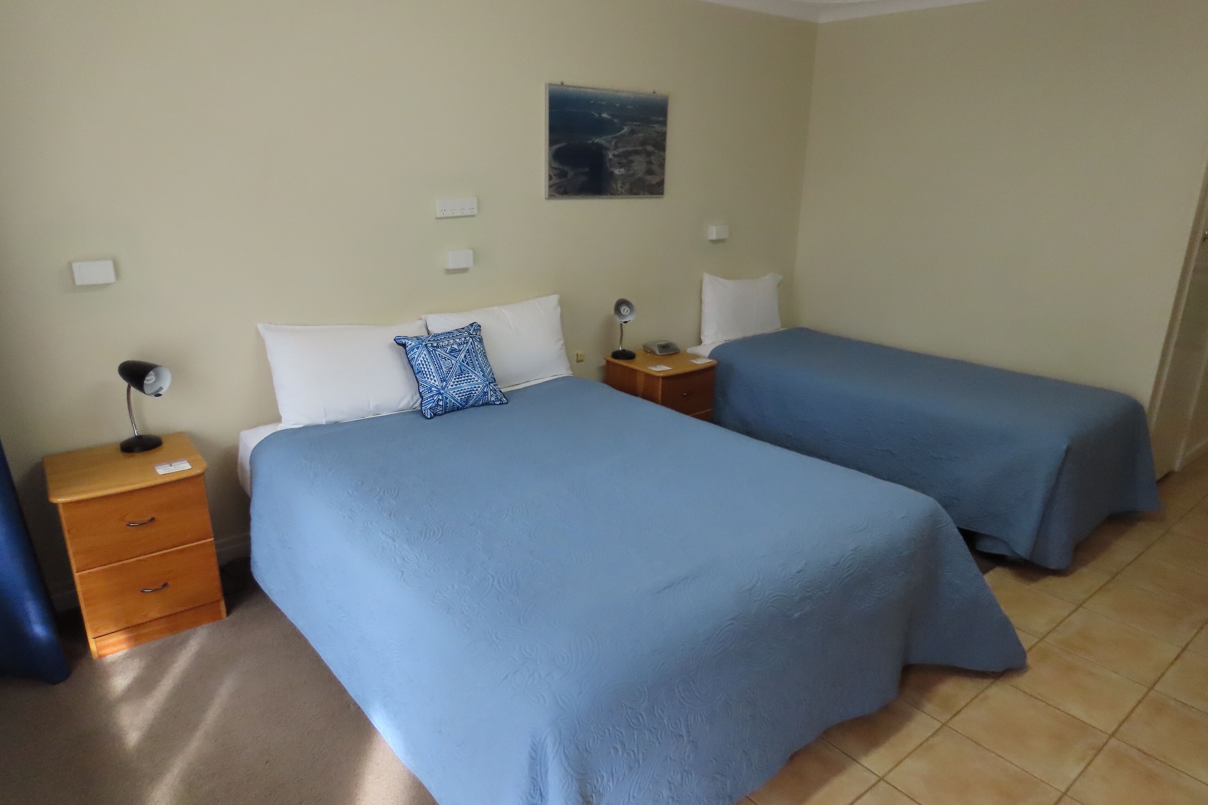 A hotel room with two beds, a wooden nightstand, a lamp, and a painting on the wall.