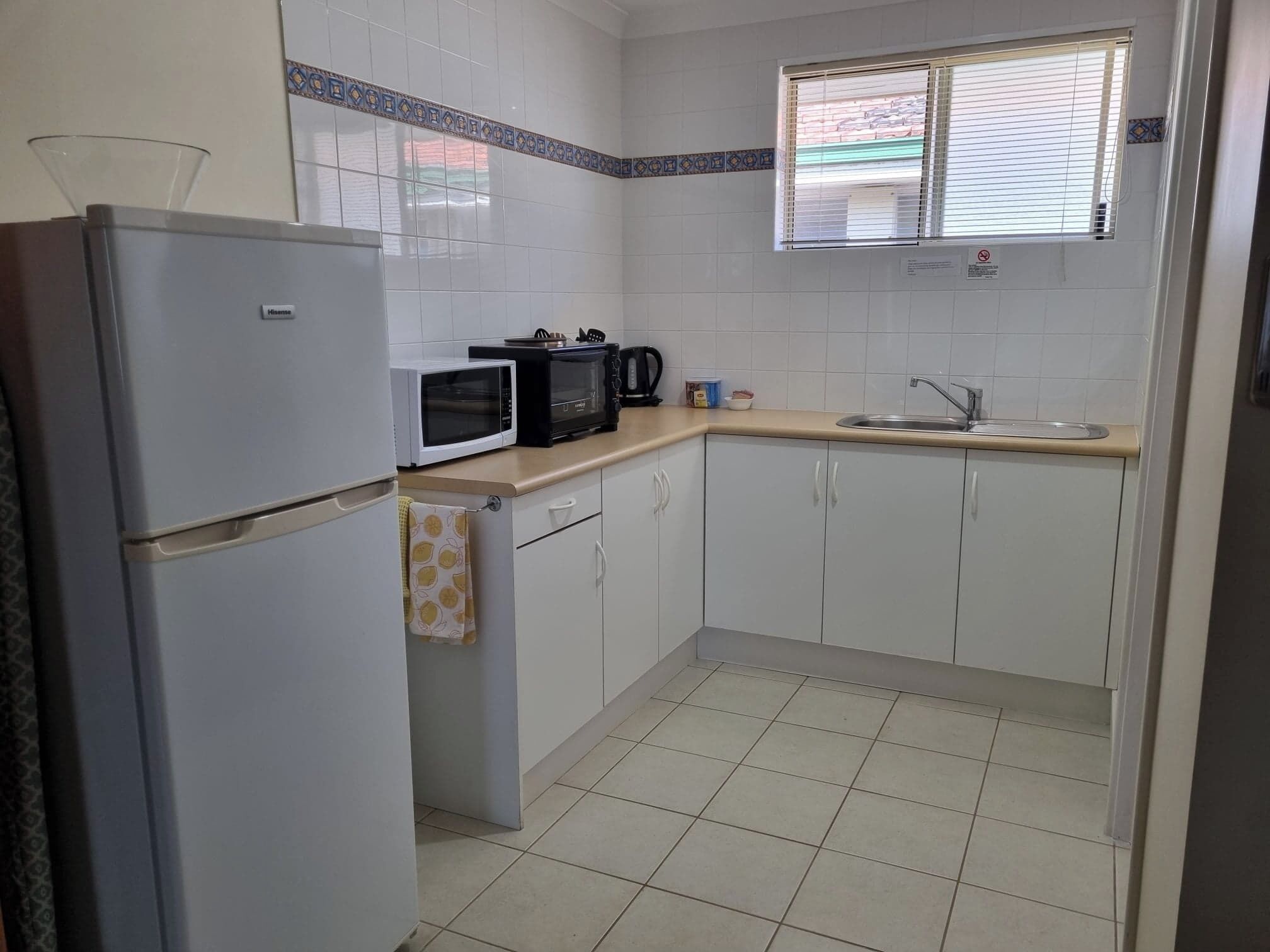 A compact kitchen with white cabinets, a refrigerator, a microwave, a coffee maker, and a window with blinds.