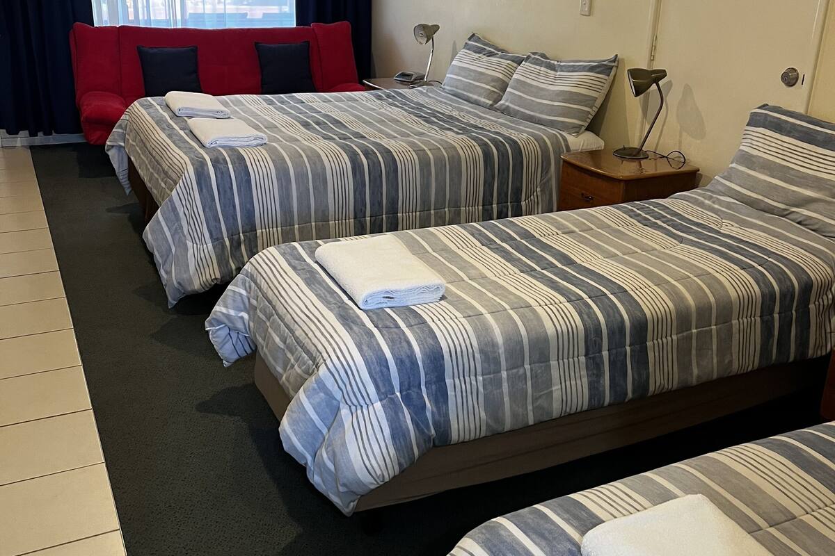 A hotel room with two beds, a red chair, a desk with a lamp, and a window with curtains.