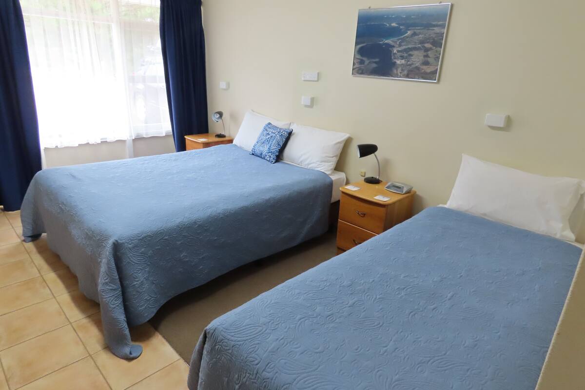 A hotel room with two beds, each with a blue bedspread, a nightstand with a lamp, and a wall-mounted picture.