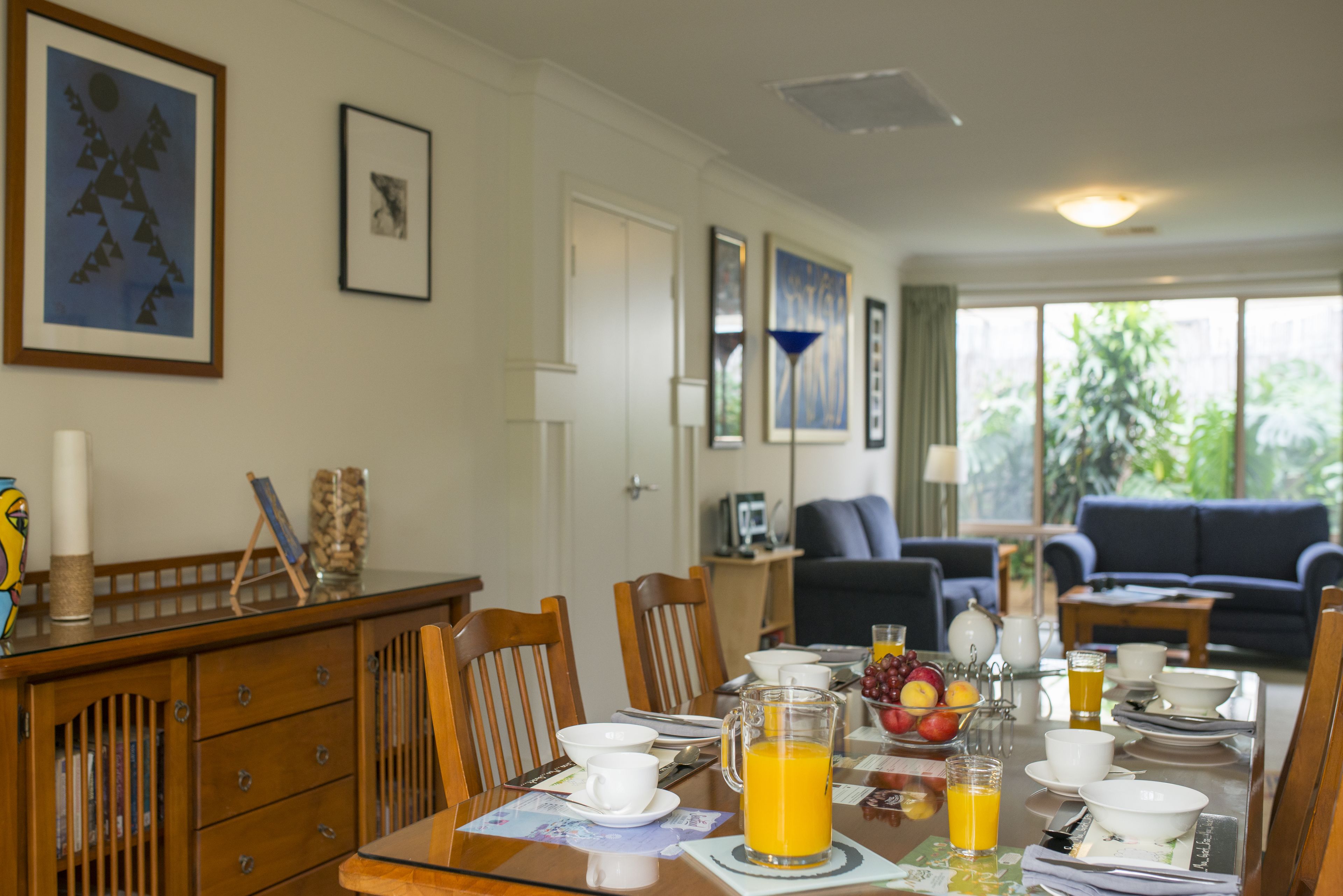 A dining area with a table set for breakfast, including orange juice, fruits, and a pitcher. There are chairs, a wooden cabinet, and framed artwork on the walls.
