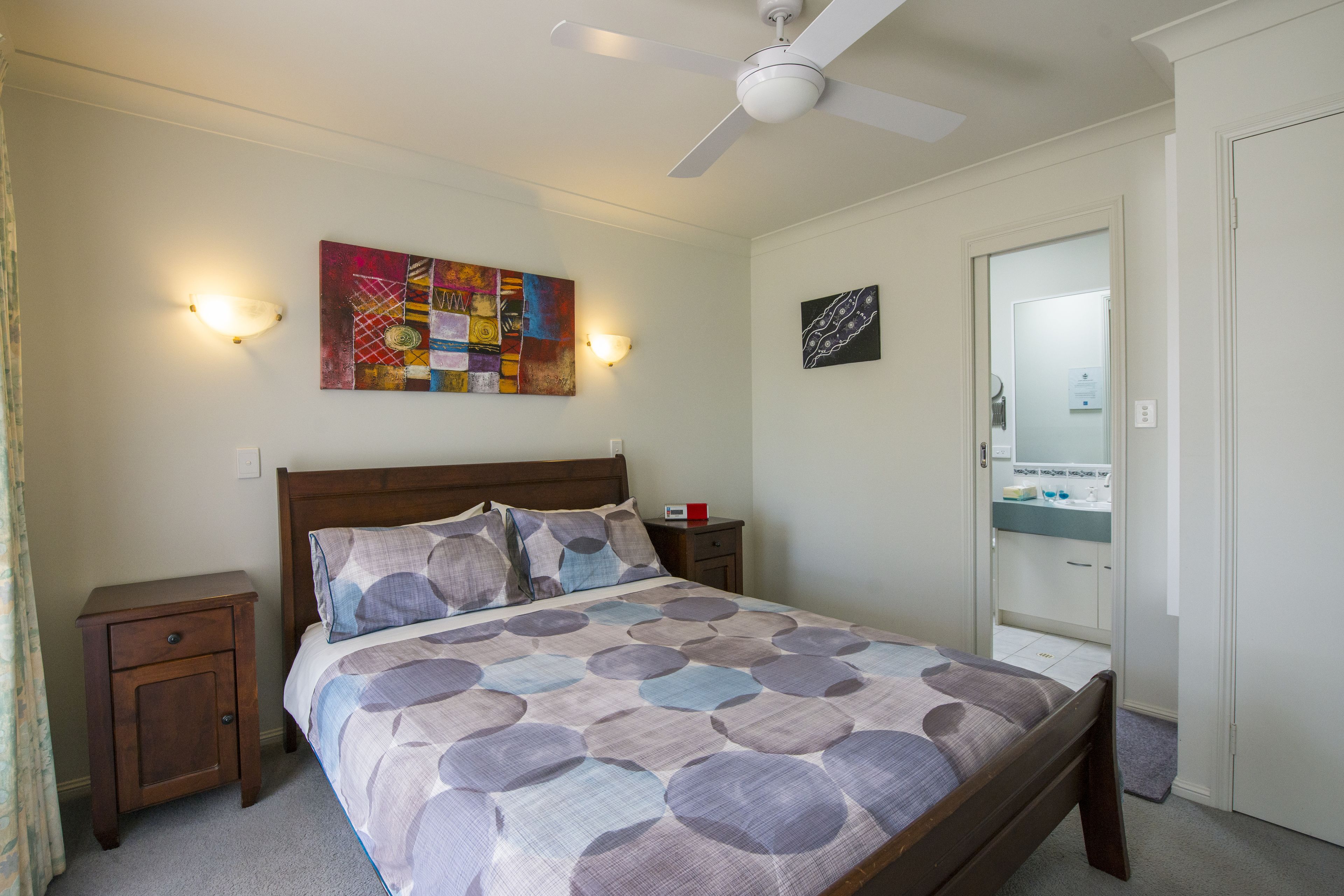 A bedroom with a bed, a nightstand, a ceiling fan, a wall-mounted light, a bathroom mirror, and a colorful artwork on the wall.