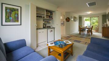A living room with blue sofas, a wooden coffee table, and a kitchen area with shelves and a refrigerator.