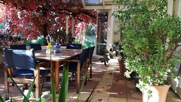 An outdoor dining area with a wooden table and chairs, surrounded by potted plants and a pergola with red foliage.