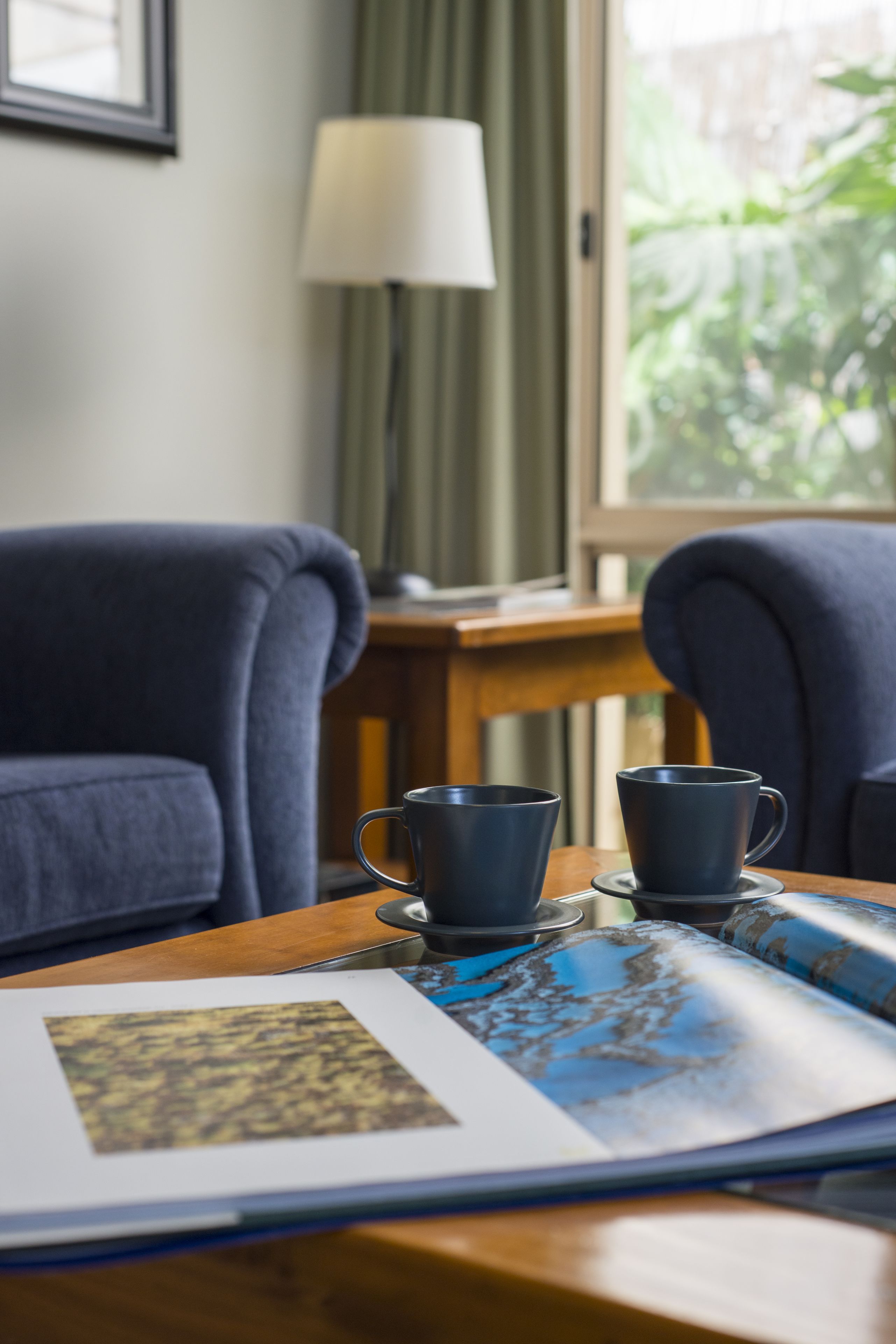 Una habitación con dos sillones azul oscuro, una mesa de madera con dos tazas azul oscuro, un cuadro enmarcado y una ventana que da a zonas verdes.