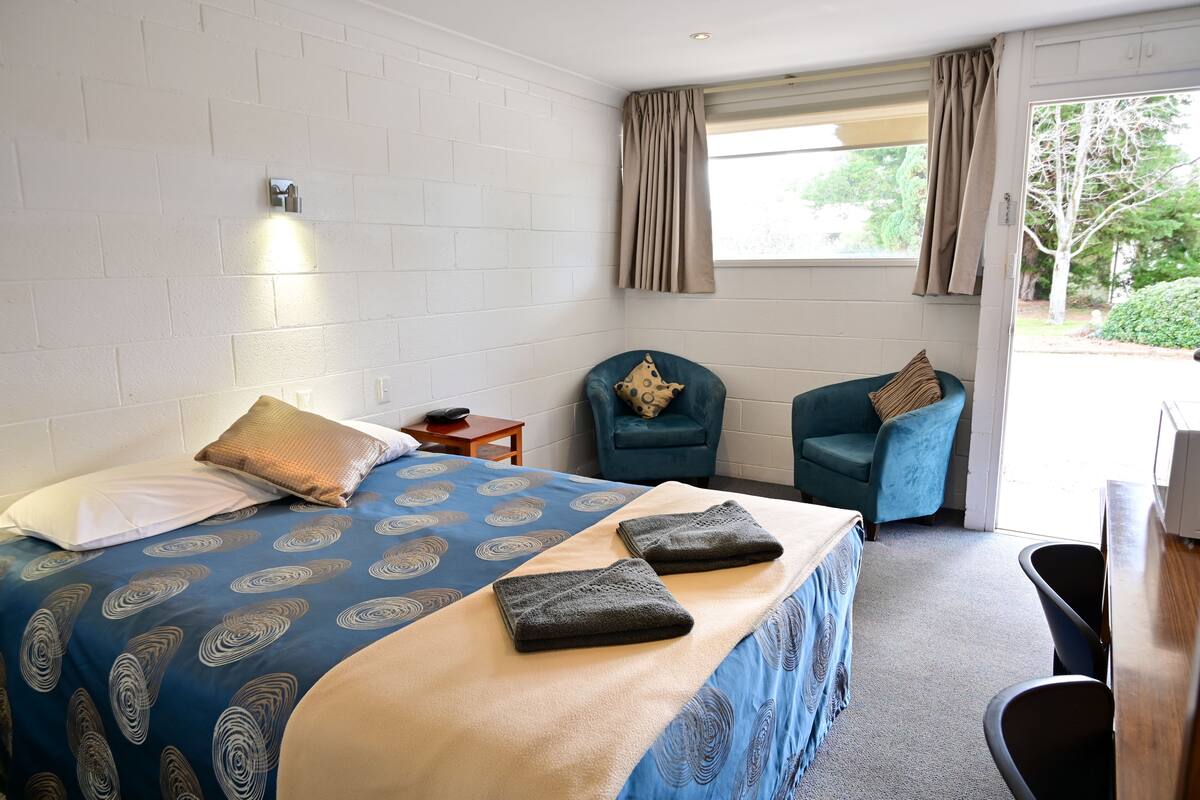 A hotel room with a bed, two chairs, a small table, and a window with curtains.