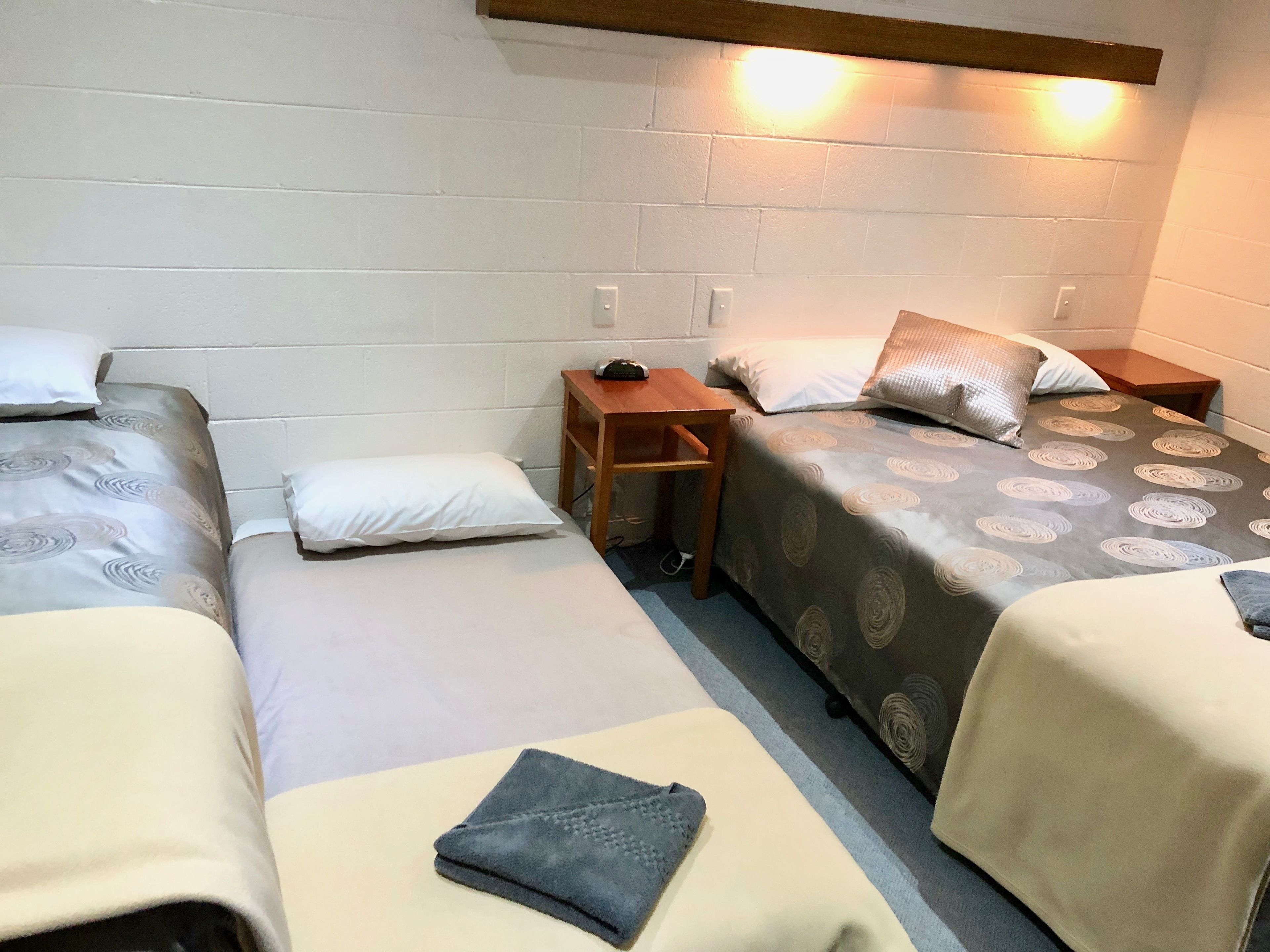 A dormitory room with three beds, each with a pillow and a blanket. There is a wooden nightstand between the beds, and a wall-mounted light above the bed on the right.